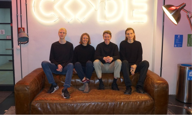 Foto von vier Personen, die auf der Rückenlehne eines Sofas sitzen. Über ihnen in Neon-Röhren leuchtend "Code", dabei besteht das "o" aus <>.